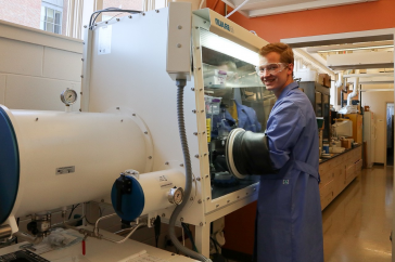 Graduate student wearing protective clothing works with his hands in a large piece of scientific equipment.